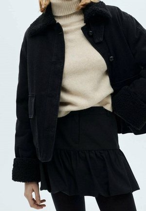 Mujer con un jersey de cuello alto beige, chaqueta negra con cuello de piel, falda negra con volante en el bajo y medias negras sobre un fondo claro.