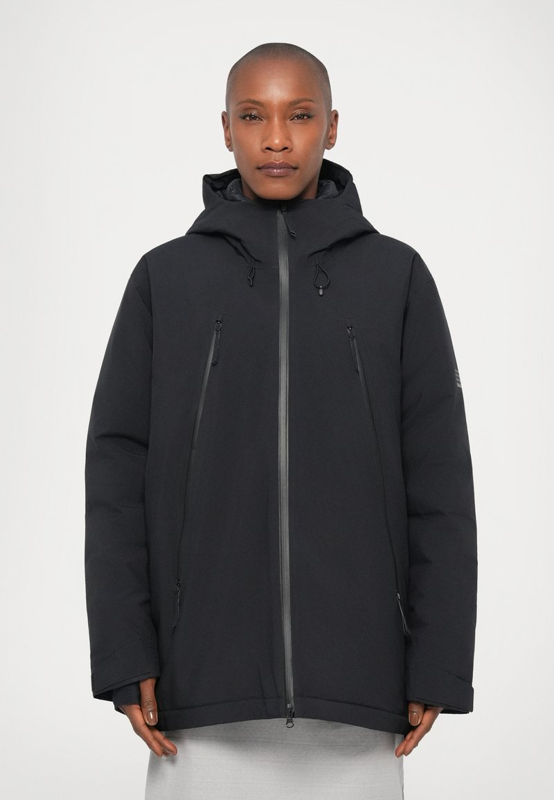 Personne portant une veste d'hiver à capuche noire avec une fermeture éclair sur la poitrine et des poches latérales, se tenant devant un fond blanc uni.