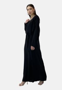 Zwarte jurk met lange mouwen van lichtgewicht stof, voorzien van een strik rond de taille en een ontspannen pasvorm, gecombineerd met witte sandalen met bandjes.