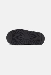 Sola de sapato preta com um padrão de pisada geométrico apresentando o logo UGG no centro, sobre um fundo branco.