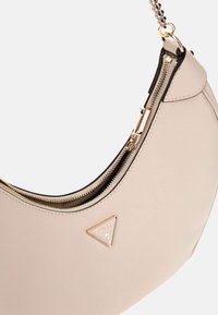 Sac à main en cuir beige avec une forme semi-circulaire, doté d'une fermeture éclair dorée et d'une plaque logo triangulaire. Surface texturée avec une finition lisse.