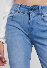 Helles blaues Denim-Jeans mit hoher Taille, Fünf-Taschen-Design und metallischem Knopfverschluss. Der Stoff zeigt subtile Auswaschungen und Textur.