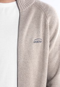 Cardigan tricoté beige clair avec un col côtelé et une fermeture éclair à l'avant. Présente un petit logo marine brodé sur la poitrine. Texture douce.