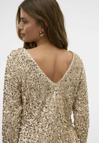 Sequinų suknelė su V formos nugarėle, turinti neutralią dramblio kaulo spalvą, ilgomis rankovėmis ir tekstūruotu paviršiumi, dekoruotu auksiniais ir sidabriniais sequinais.