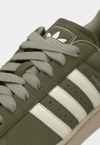 Sneakers in pelle scamosciata verde con lacci grigi, caratterizzate da tre strisce bianche sul lato, una toppa con logo sulla lingua e una punta rinforzata.