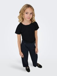 Enfant blonde aux cheveux bouclés portant un haut noir à manches courtes, un pantalon large bleu marine et des mocassins noirs, debout devant un fond blanc uni.