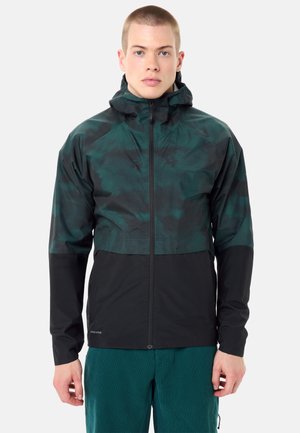 Jonge man draagt een zwarte en donkergroene zip-up capuchonjas en teal broek, staand tegen een eenvoudige lichte achtergrond.