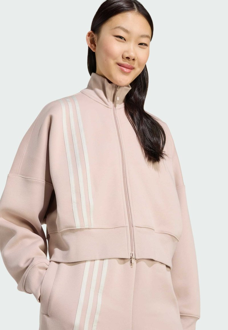 Lyserød cropped zip-up sweatshirt lavet af blødt stof, med tre hvide lodrette striber på venstre skulder og en høj hals.