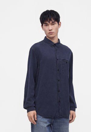 Jeune homme aux cheveux courts et foncés, portant une chemise bleu marine boutonnée avec une poche poitrine et un jean bleu, debout sur fond blanc.
