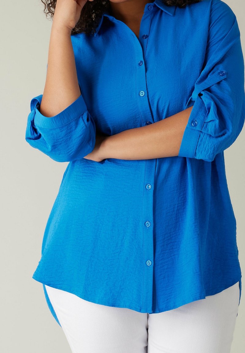 Person med lockigt hår som bär en ljusblå långärmad skjorta med uppkavlade ärmar och vita byxor, med armarna i kors.