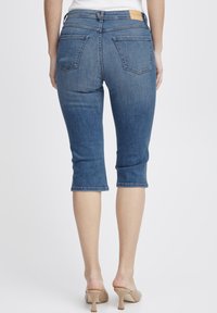 Denim capri hlače v srednjem modrem odtenku z petimi žepi, končanimi robovi in oznako na pasu. Nosi se z bež sandali na visoki peti.