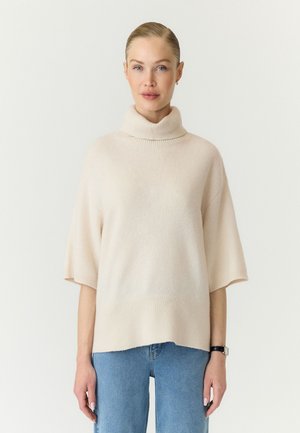 Femme à la peau claire et aux cheveux blonds, portant un pull à col roulé crème à manches courtes et un jean bleu, debout devant un fond blanc uni.