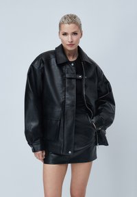 Veste en cuir noire surdimensionnée avec fermeture à bouton, poches avant et col structuré. Texture lisse et finition mate.