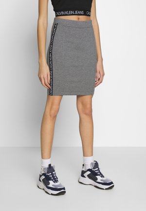 Femme portant une jupe grise mi-genoux avec une bande verticale du logo Calvin Klein Jeans, un crop top noir, des chaussettes blanches et des baskets noir et blanc.