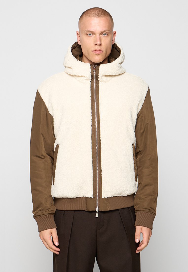 Homme portant une veste zippée marron avec un devant et une capuche en sherpa beige, se tenant contre un fond gris clair uni.