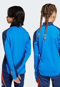 Des vestes de sport bleues présentent des manches longues et des cols hauts, avec des accents marins et une bordure orange sur les côtés, fabriquées en un tissu lisse et élastique.