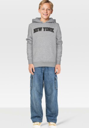 Pelēks kapučjaks ar melnu uzrakstu "NEW YORK", apvienots ar brīviem zilām džinsu kravas biksēm un baltām sporta apaviem, uz vienkārša fona.