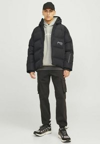 Sort pufferjakke med lynlåslommer, grå hættetrøje under, sorte cargo bukser og sorte sneakers. Model iført en sort kasket.