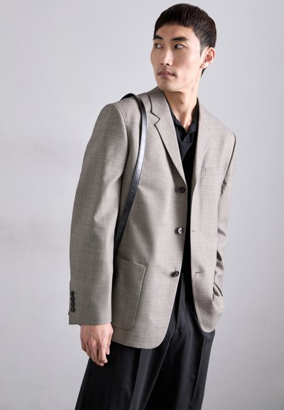 Hellgrauer, maßgeschneiderter Blazer mit Revers, drei Knöpfen, zwei Fronttaschen und strukturiertem Stoff, kombiniert mit einem schwarzen Hemd und einer Tasche.