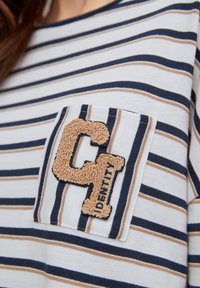 T-shirt in cotone a righe con linee orizzontali blu, crema e marrone; presenta una toppa marrone testurizzata con una "C" e "IDENTITY" ricamate sulla tasca.