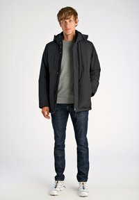 Mustat toppatakki hupulla, harmaa neulepaita, tummat farkut ja valkoiset lenkkarit värikkäillä nauhoilla, seisoo vaaleaa taustaa vasten.