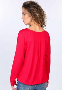 Rotes Langarmshirt mit einem Falten-Detail am Rücken und rundem Ausschnitt, aus einem weichen, leichten Stoff gefertigt. Kombiniert mit blauen Jeans.