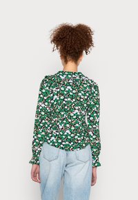 Blusa de manga longa em preto com um padrão floral verde. Apresenta ombros franzidos e punhos com franzido, combinada com jeans azul claro.