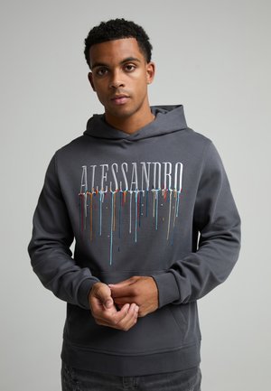 Jeune homme portant un sweat à capuche gris foncé avec le texte "ALESSANDRO" et des gouttes de peinture colorées, debout les mains légèrement croisées.