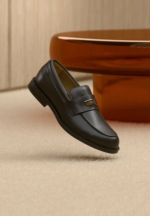 Mocasín de cuero negro con una placa metálica de marca apoyado cerca de una mesa redonda marrón sobre un suelo alfombrado beige.