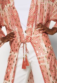 Mönstrad kimono i korall med guldaccenter, med ett knytliv och tofsar. Tillverkad av lätt tyg med vida ärmar och öppen framsida.