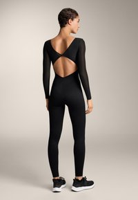 Fekete body hosszú, áttetsző ujjakkal és nyitott hátrésszel, V-alakú kivágással. A modell fekete tornacipőt visel. Sima textúra.