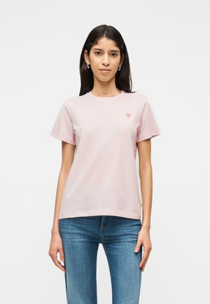 Mladá žena s dlouhými tmavými vlasy, oblečená v světle růžovém tričku s krátkými rukávy a modrých džínách, čelící dopředu na jednobarevném pozadí.