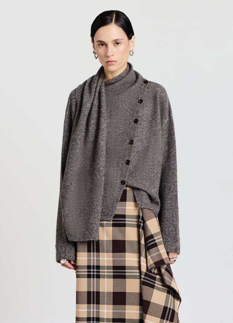 Maglione grigio lavorato a maglia con collo alto, sette bottoni scuri lungo il lato, abbinato a una gonna a quadri beige con accenti marroni e neri.