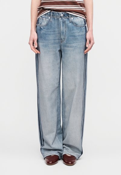 Φαρδιά τζιν σε ανοιχτό γαλάζιο με ξεθωριασμένο πλύσιμο, δύο μπροστινές τσέπες και πλευρικές ρίγες σε πιο σκούρο μπλε. Συνδυασμένα με καφέ παπούτσια και ριγέ μπλούζα.