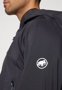 Dunkelgraue Jacke mit Kapuze, ausgestattet mit einem Reißverschluss und einem weißen Logo eines Mammuts auf dem linken Ärmel. Glatte Textur und leichtes Design.