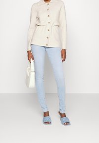Cárdigan de punto beige con cuello y bolsillos frontales, combinado con jeans ajustados de color azul claro y sandalias de tacón rizadas azules. Bolso blanco.