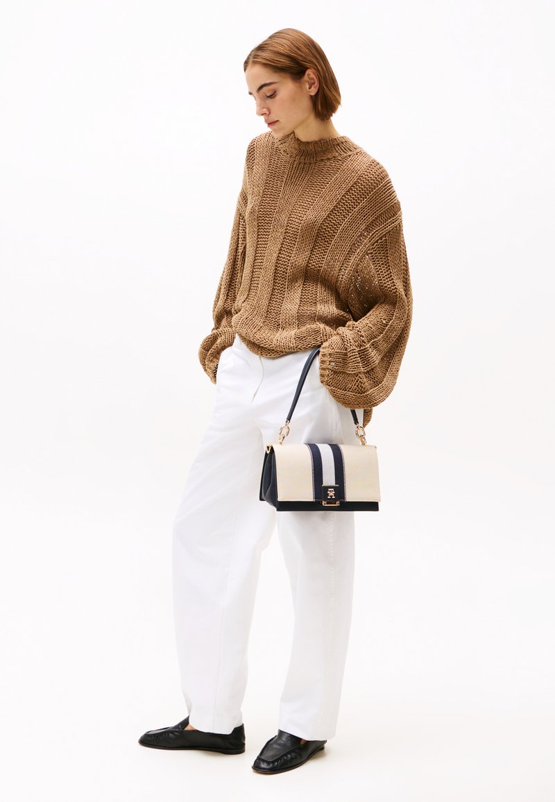 Brauner Pullover aus Strick mit Rippstruktur, weiße Hose, schwarze Slipper und eine zweifarbige Tasche mit creme- und marinefarbenen Streifen sowie goldener Hardware.