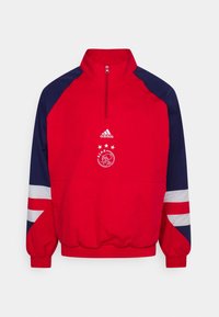 Cazadora roja con mangas marinas, cuello con media cremallera, puños y cinturilla elásticos. Presenta el logo de Adidas y el emblema del Ajax con estrellas.