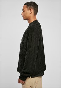 Svart strikket cardigan med en strukturert kabelmønster, med rund hals og knappelukking, vist fra siden.