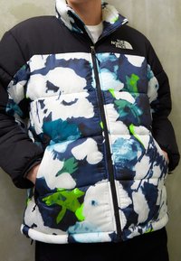 Cazadora tipo puffer con un patrón floral en azul marino, blanco y verde. La mitad superior es de color negro sólido, con cierre de cremallera y bolsillos laterales.