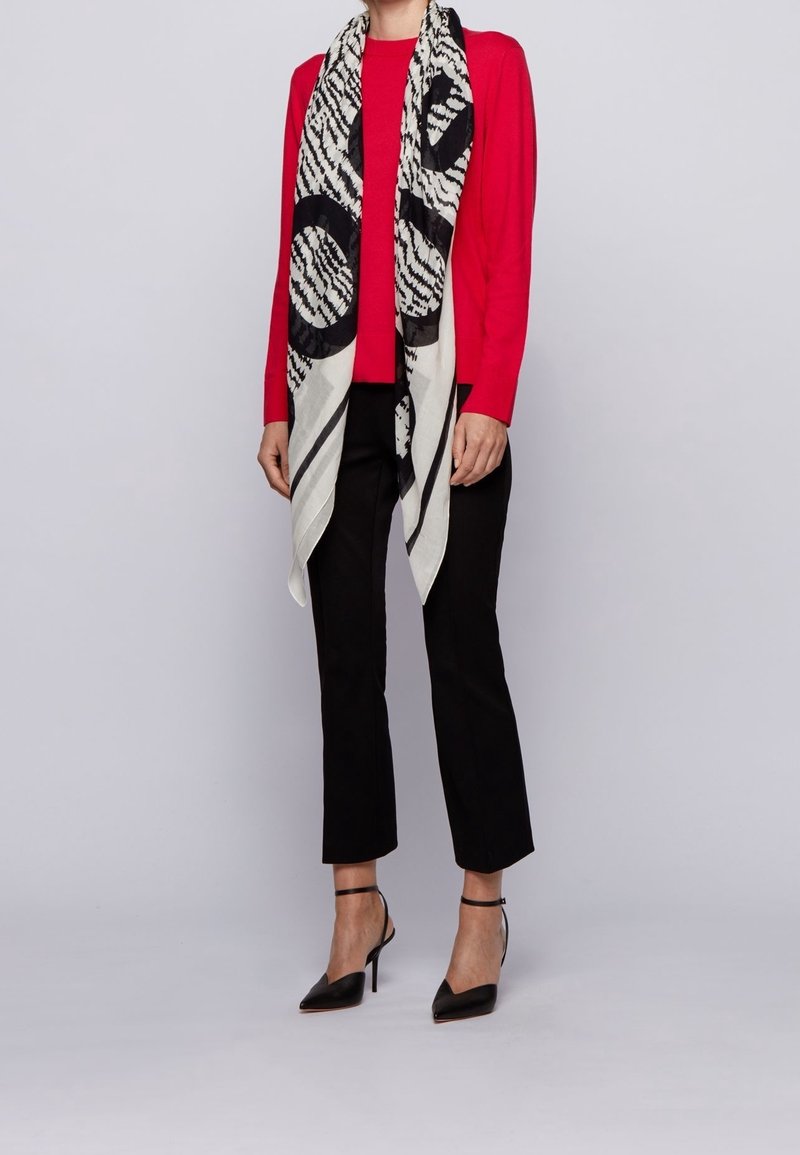 Femme portant un haut rouge à manches longues, un pantalon noir, des talons hauts noirs et une longue écharpe noire et blanche à motifs drapée autour du cou.