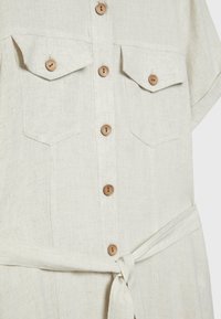 Hellbeige Leinenhemdkleid mit kurzen Ärmeln, zwei Brusttaschen, Holzknöpfen und einem gebundenen Taillengürtel. Texturiertes Stoff mit einem subtilen Muster.