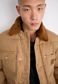 Braune gepolsterte Jacke mit einem sandfarbenen Kunstfellkragen, goldenen Beschlägen und Fronttaschen. Über einem schwarzen Hemd mit drei Knöpfen getragen.