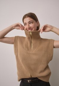 Light tan knitted sweater with a high zip collar, short sleeves, and a loose fit. Textured with a breathable open weave pattern.