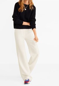 Czarny sweter dziergany na drutach, białe spodnie o szerokich nogawkach oraz wielokolorowe trampki. Naszyjnik z okrągłą zawieszką. Skupienie na fakturze i kontraście kolorów.