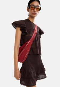 Brązowa sukienka warstwowa z falbaniastymi rękawami, zestawiona z czerwoną torebką crossbody z srebrnymi ćwiekami. Modelka nosi duże przyciemniane okulary przeciwsłoneczne.