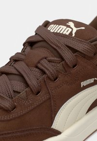 Gros plan sur une basket Puma marron avec des lacets épais, une texture en daim, un logo Puma blanc et une bande latérale de couleur crème.