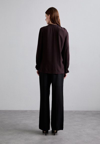 Blusa a maniche lunghe marrone scuro con polsini arricciati, abbinata a pantaloni neri a gamba larga con vita alta e tacchi neri. Trame di tessuto lisce.