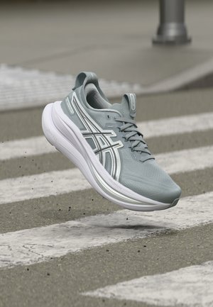 Chaussure de course gris clair avec semelle blanche en l'air au-dessus d'un passage pour piétons en asphalte avec des particules de gravier dispersées.