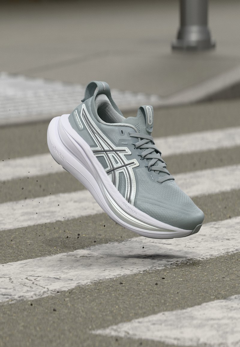 Γκρι αθλητικό παπούτσι με λευκή σόλα στον αέρα πάνω από διαβάτη πεζών σε άσφαλτο, με διασκορπισμένα κόκκους χαλικιού.